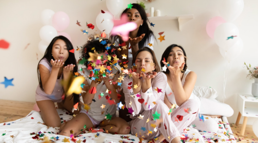 A group of women are sitting on a bed blowing confetti and balloons.