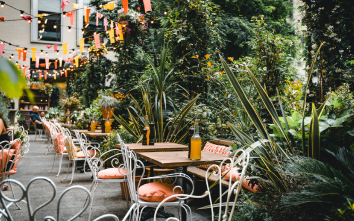 A row of tables and chairs in a garden surrounded by trees.