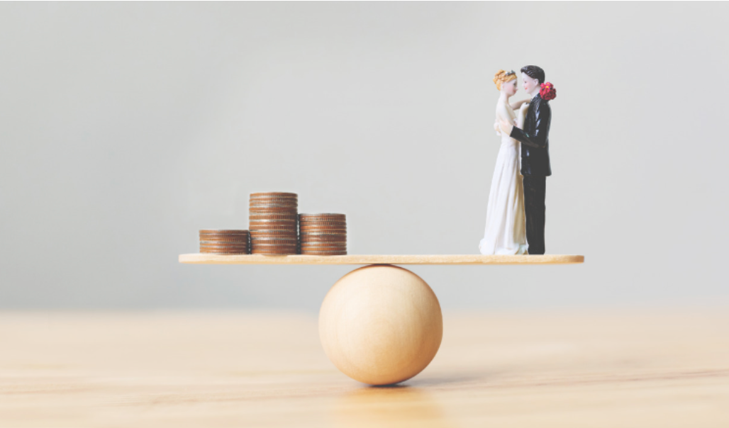 A bride and groom figurine are standing on a balance beam with stacks of coins.