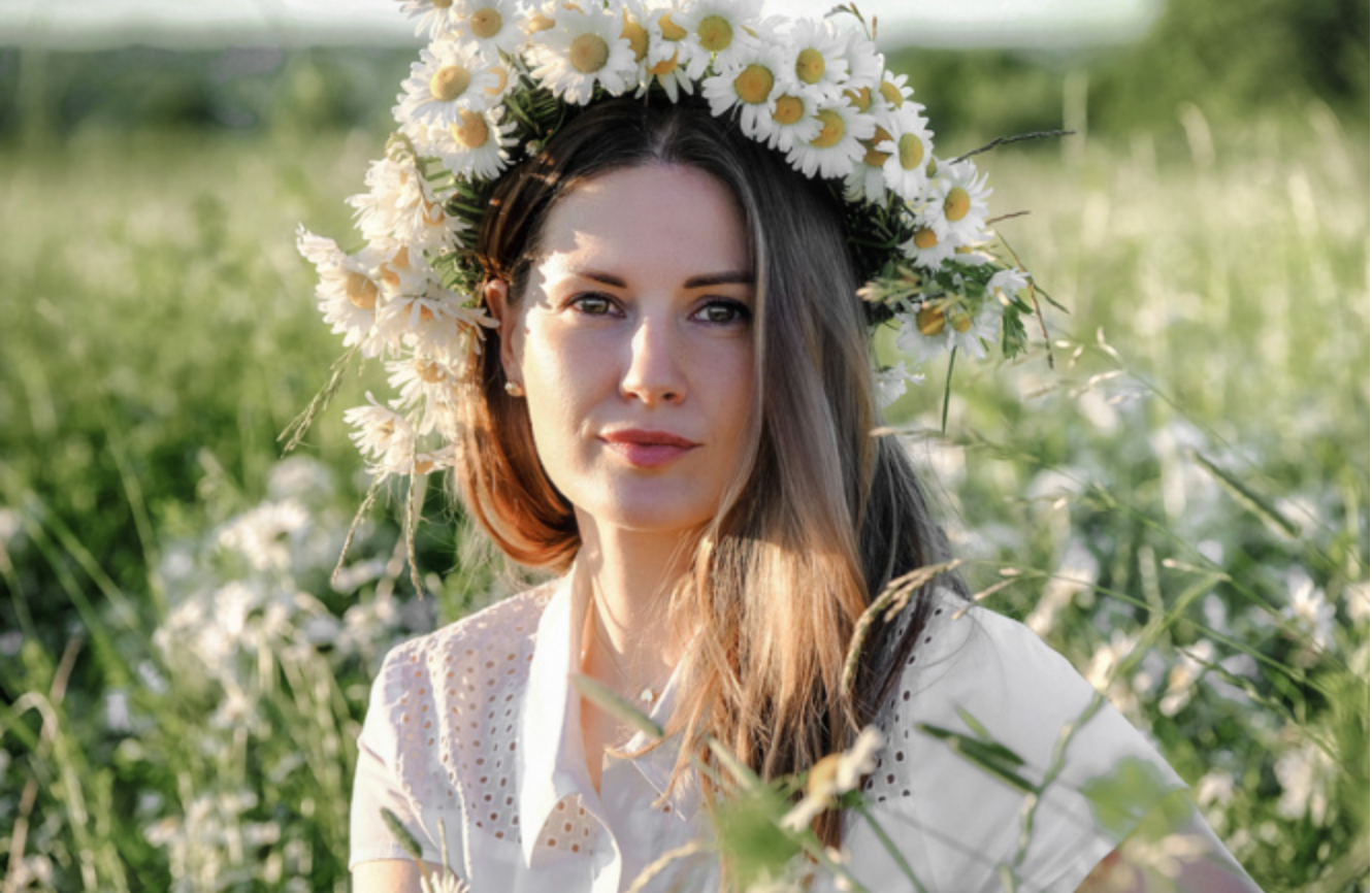 A woman wearing a wreath of daisies on her head is sitting in a field of daisies.