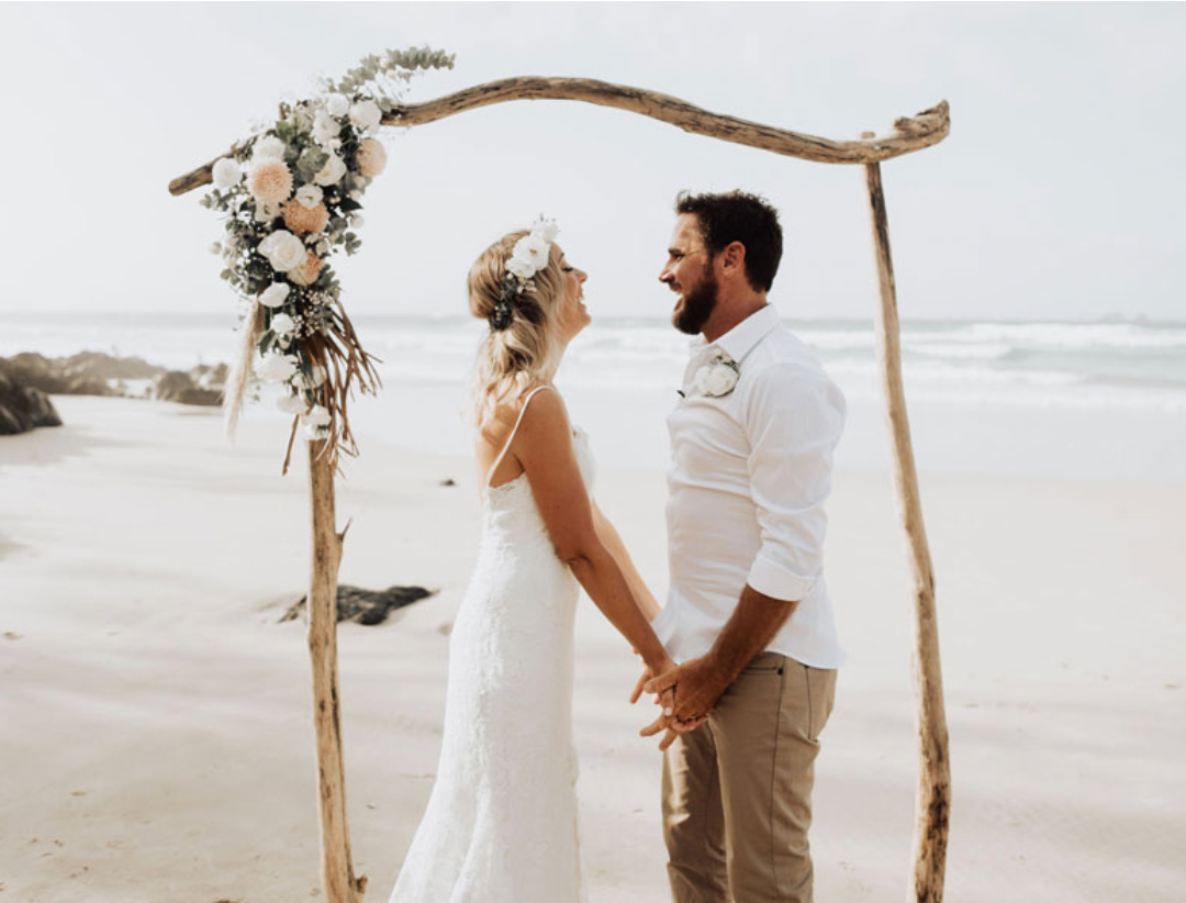 A bride and groom are holding hands on the beach under a wooden arch.