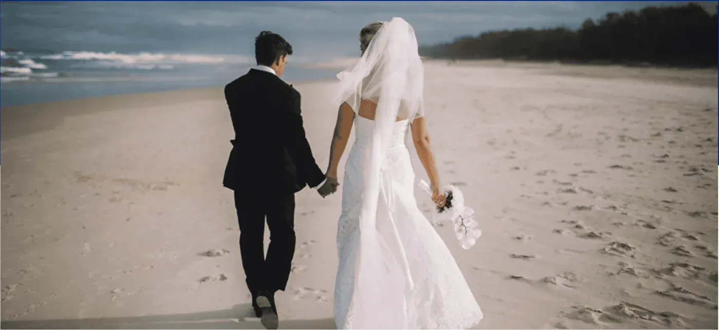 A bride and groom are standing on a beach holding hands.