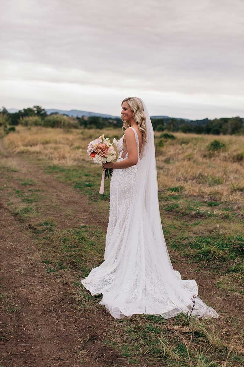 A bride in a white dress and veil is standing in a field holding a bouquet of flowers.
