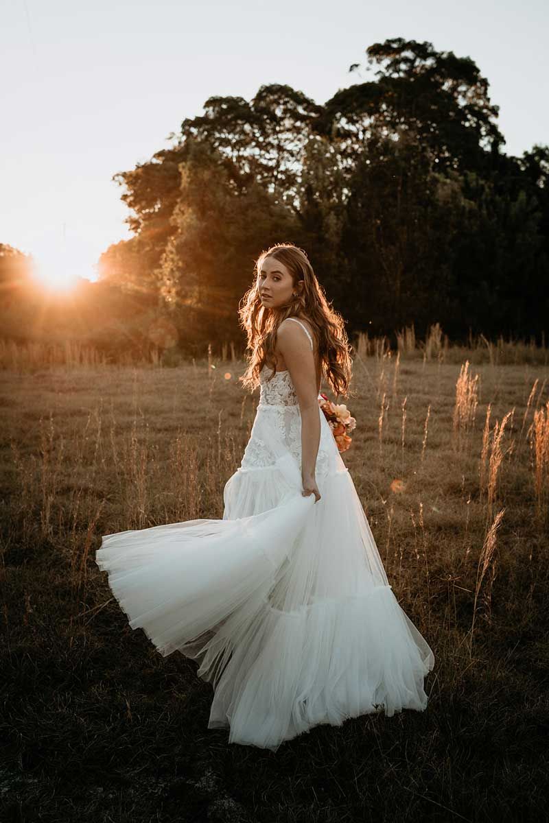 A woman in a wedding dress is standing in a field at sunset.