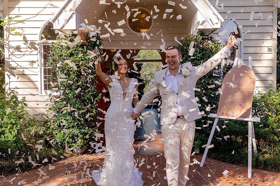 A bride and groom are standing in front of a building while confetti is being thrown at them.