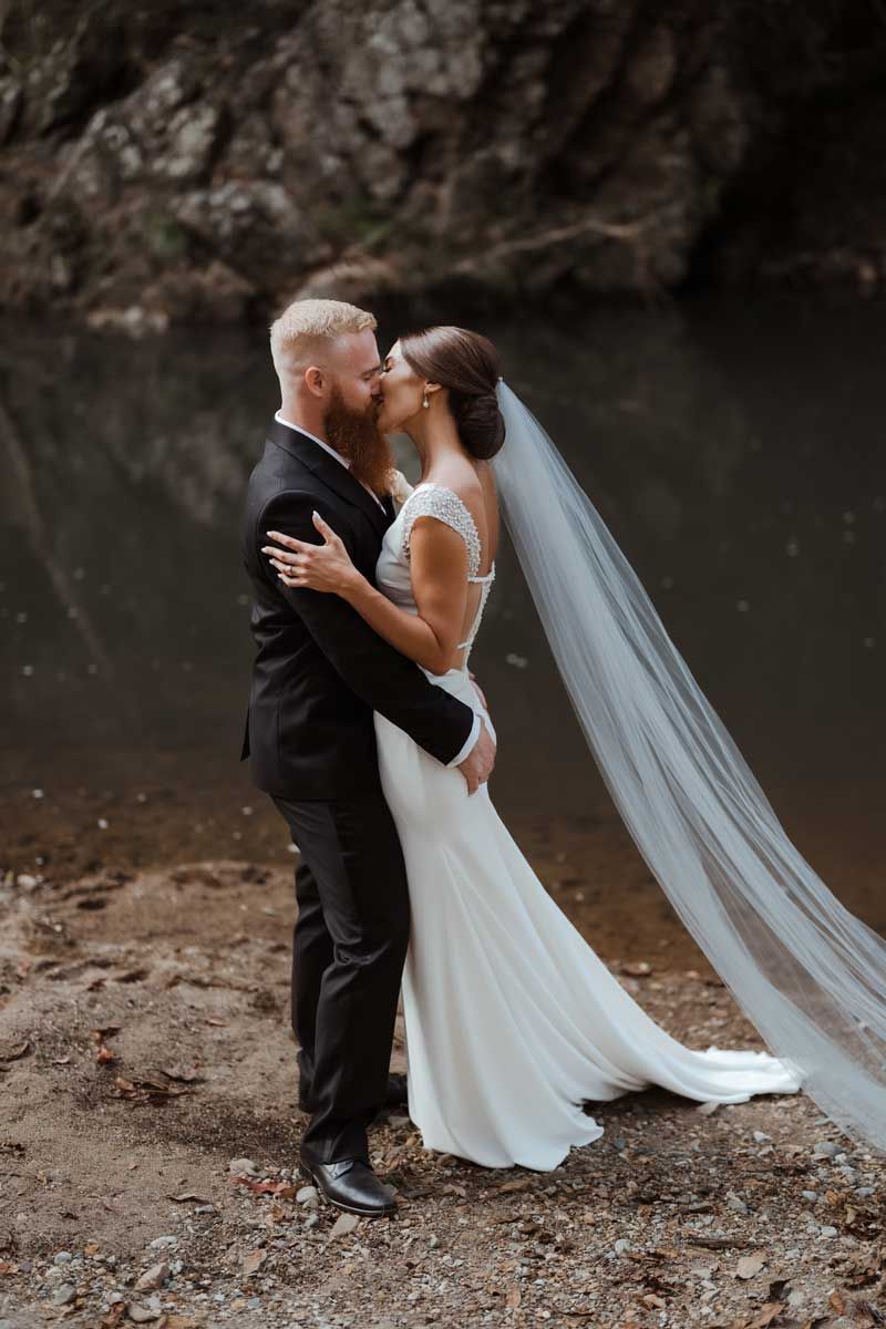 A bride and groom are kissing in front of a body of water.