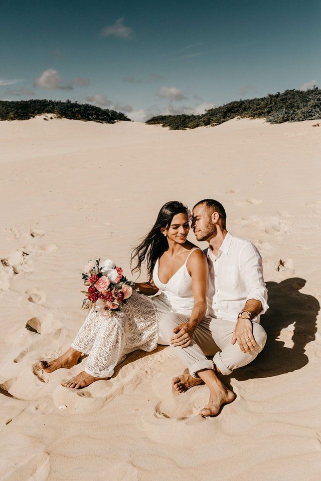 A bride and groom are sitting on top of a sand dune.