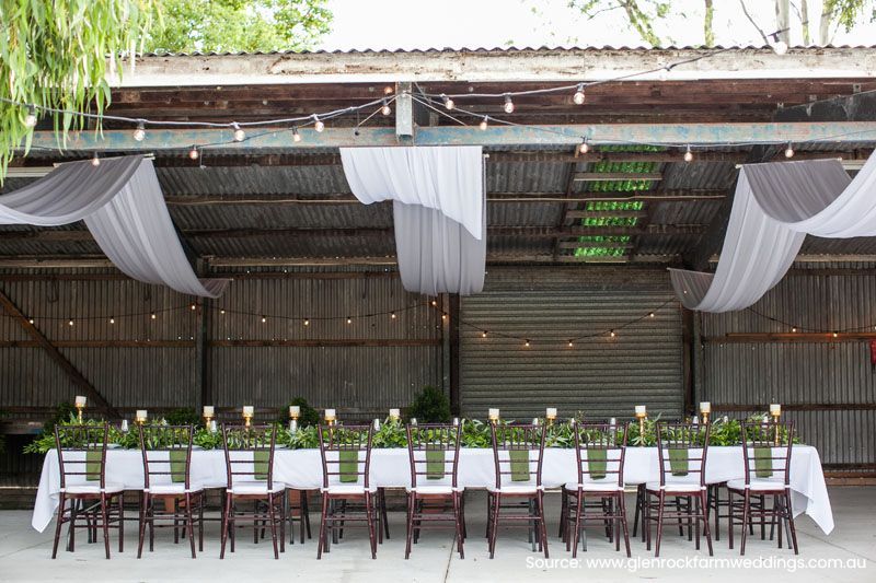 A long table with white tables cloths and chairs in a barn