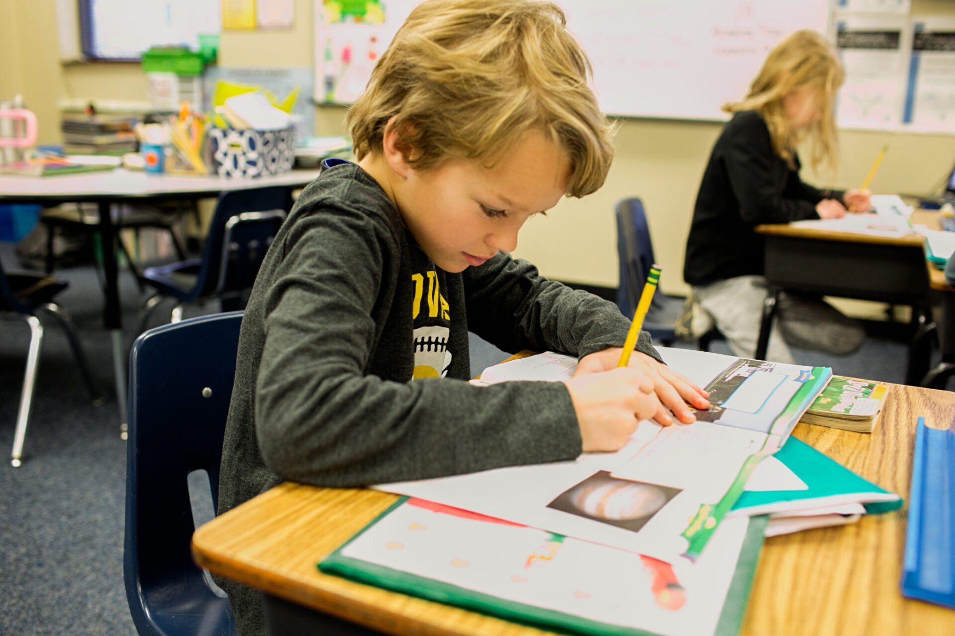 A young boy is sitting at a desk in a classroom writing in a notebook.