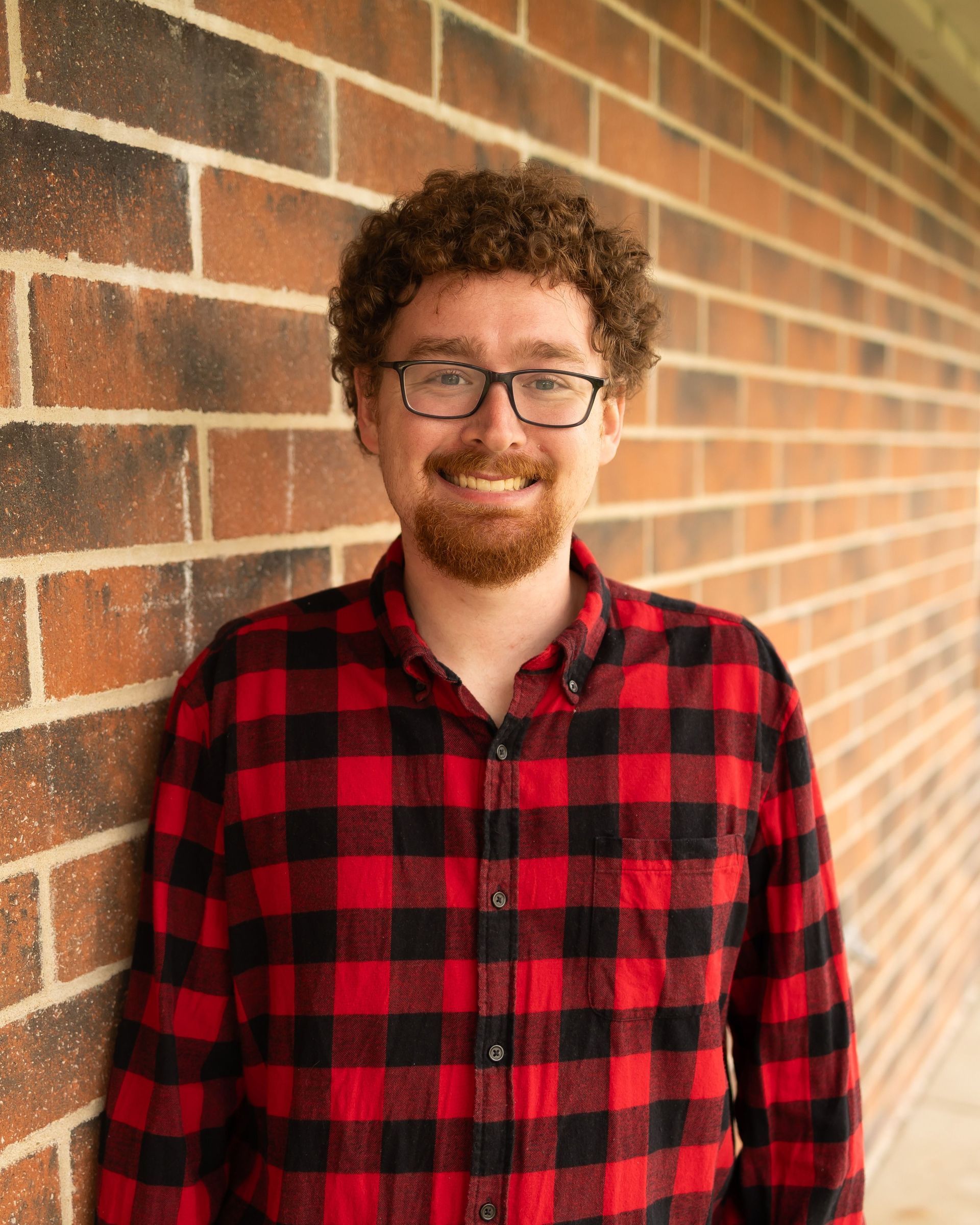 A man wearing glasses and a tan shirt is smiling in front of a brick wall.