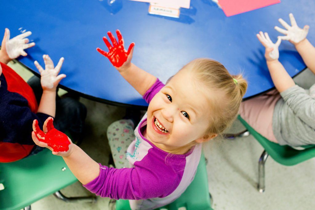 A little girl is sitting at a table with her hands painted red.