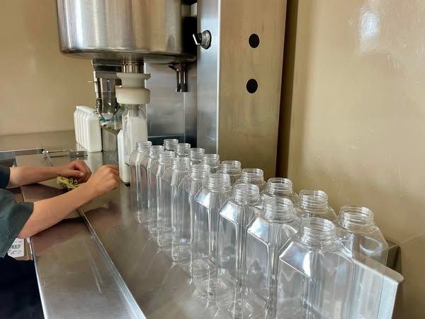 A worker fills empty clear plastic bottles using an automated liquid filling machine on a stainless steel counter.