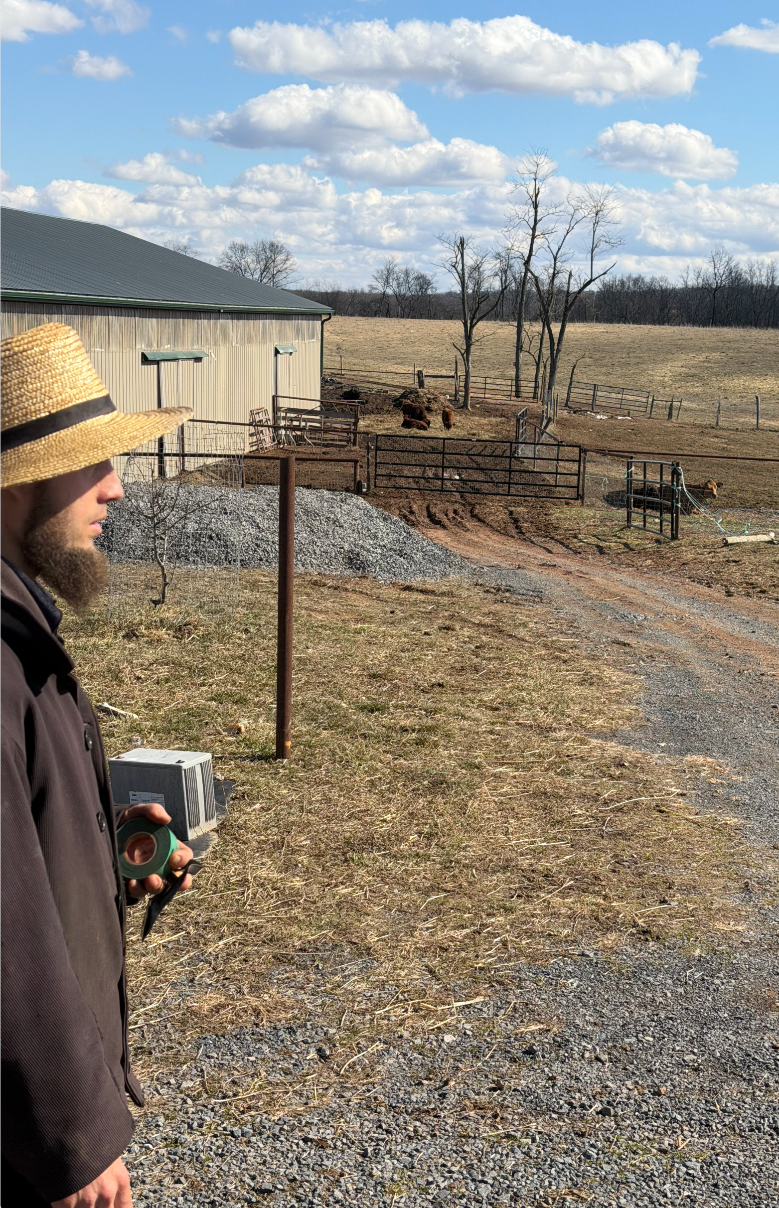 A person in a straw hat stands outdoors near a long metal barn overlooking a fenced, dirt-filled animal enclosure.