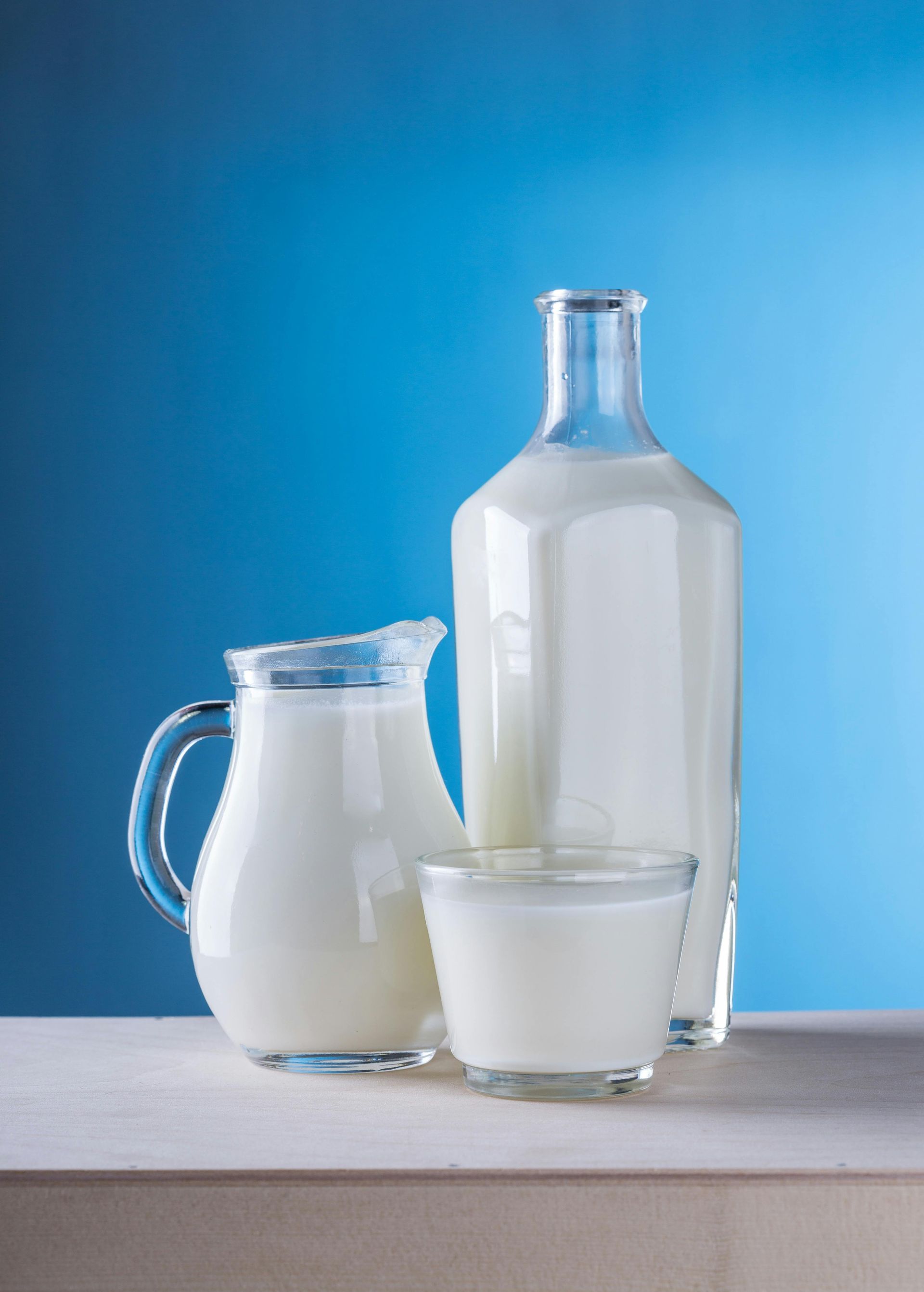 A glass pitcher, a tall bottle, and a glass filled with milk on a table against a solid blue background.