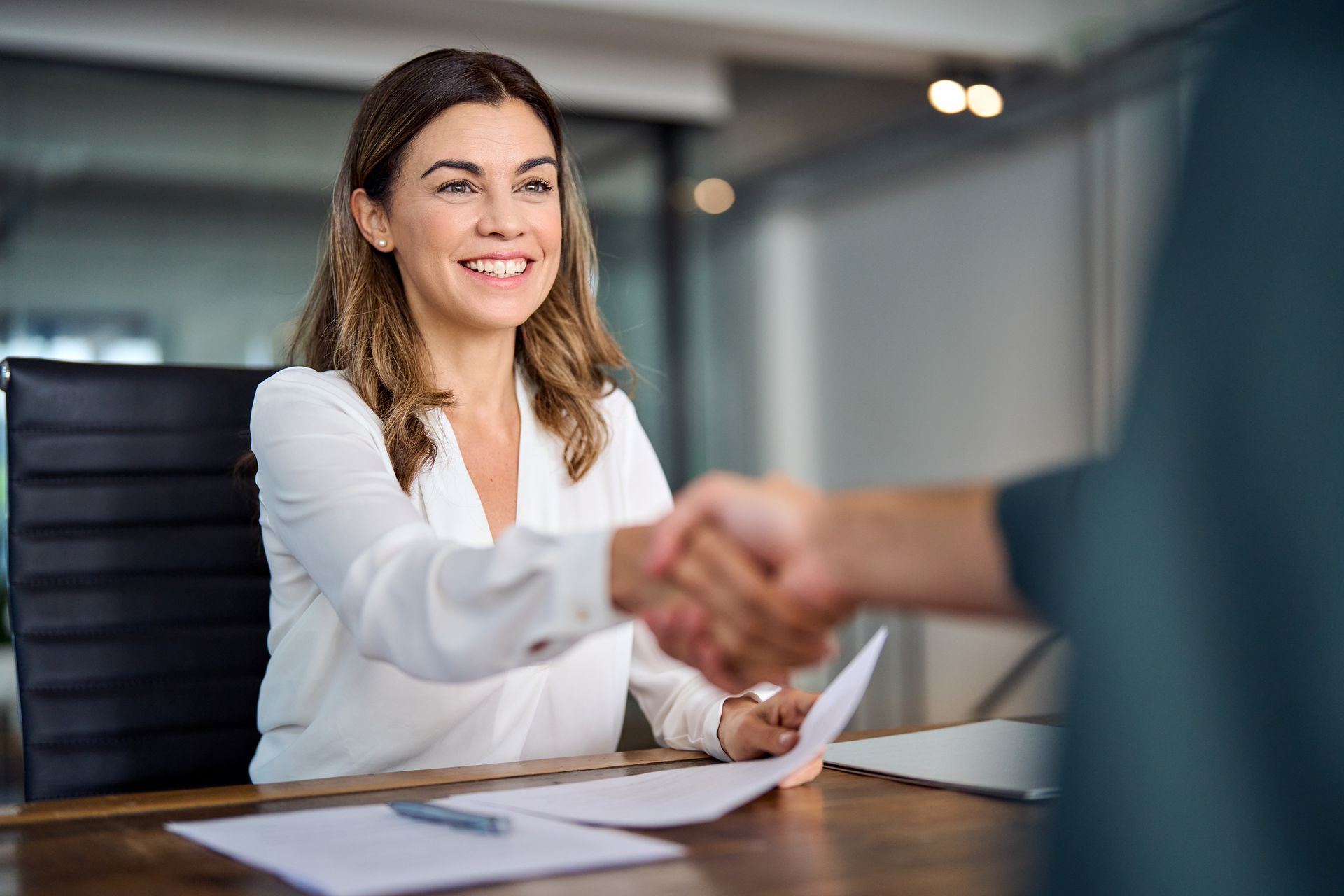 A professional shaking hands with a client across a desk with documents and a pen laid out.