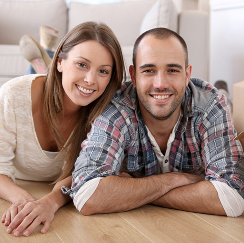 Couple smiling, lying on floor, near couch. Man in flannel shirt, woman in white top.