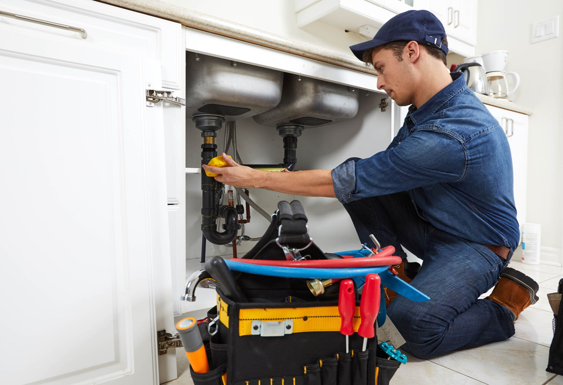 Plumber kneeling under a kitchen sink, working on pipes. He wears a blue cap and denim shirt, and his toolbox is open.