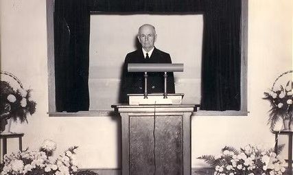 Man standing behind a podium on a stage with floral arrangements and curtains