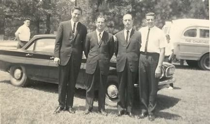 Four men standing beside a vintage car in a tree-lined outdoor setting.