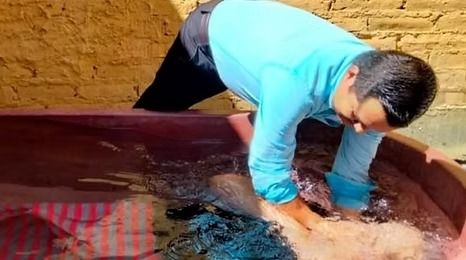Person in a blue shirt leans over a red basin, washing fabric in water against a brick wall.