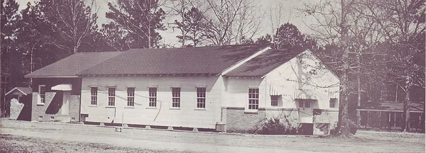 Sepia photo of a small rural schoolhouse with people standing outside and trees in the background