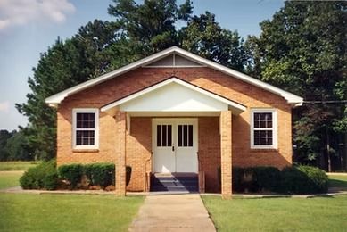 Small brick building with white trim and central porch, fronted by a walkway and lawn.