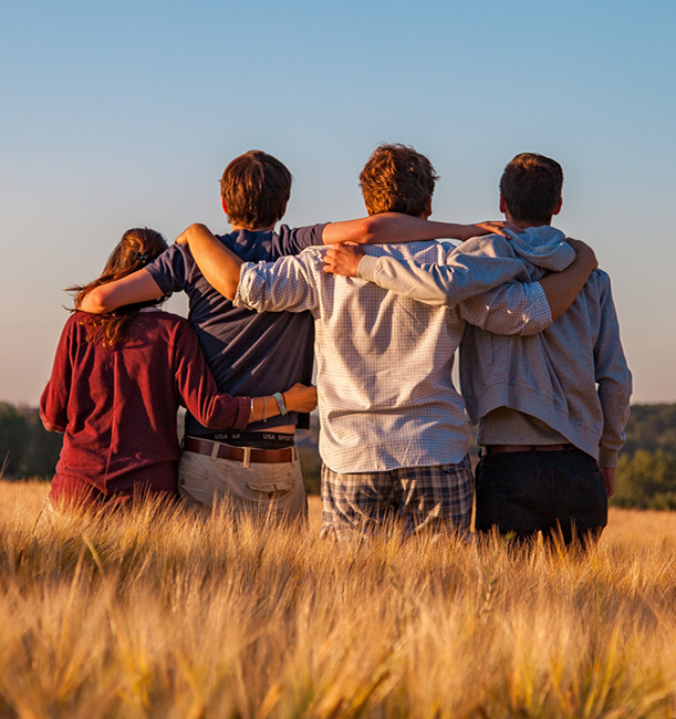 Four friends with arms around each other stand in a wheat field, looking out at a sunset.