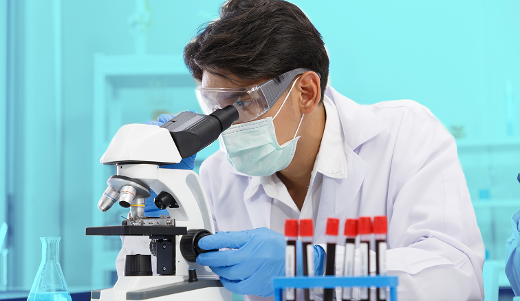 Scientist in lab coat, mask, and goggles examines a sample under a microscope, surrounded by lab equipment.