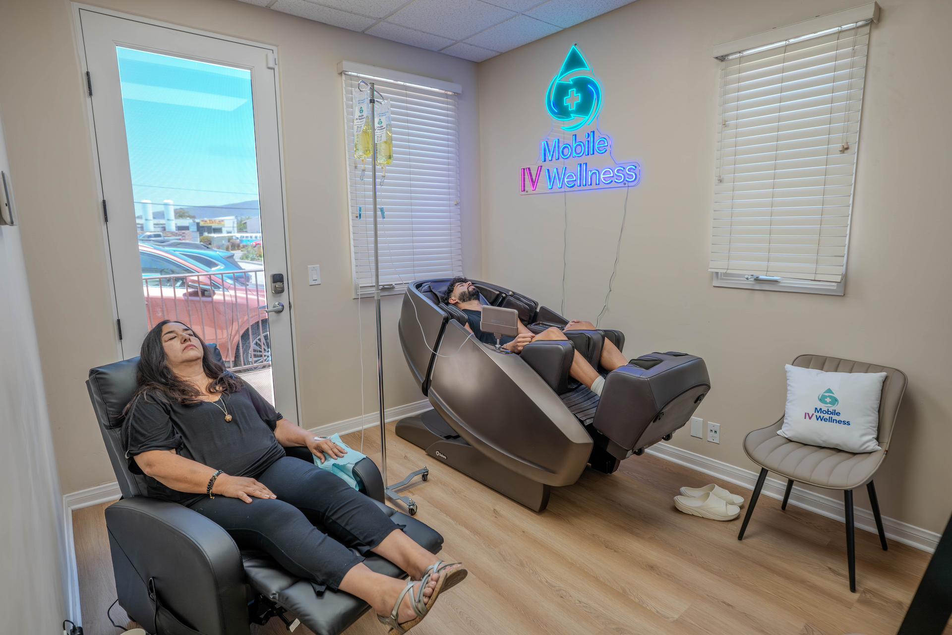 Woman in a white robe sits in a chair, receiving an IV drip. Reading a tablet, room with white tile wall.