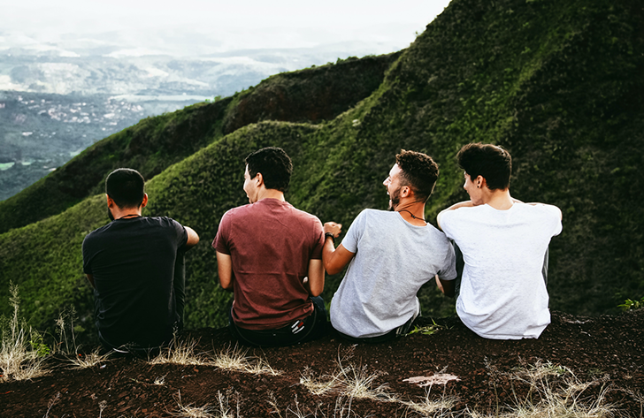 Four young men sit on a hillside, overlooking a valley; one smiles, others look at the view.