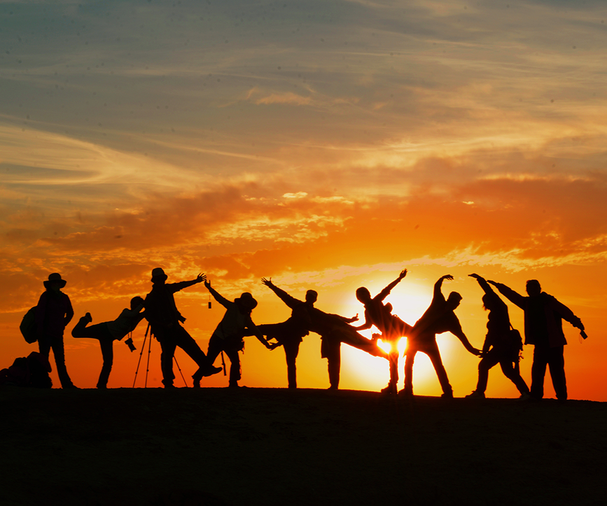 Silhouetted group of people posing playfully on a hilltop against a vibrant sunset.