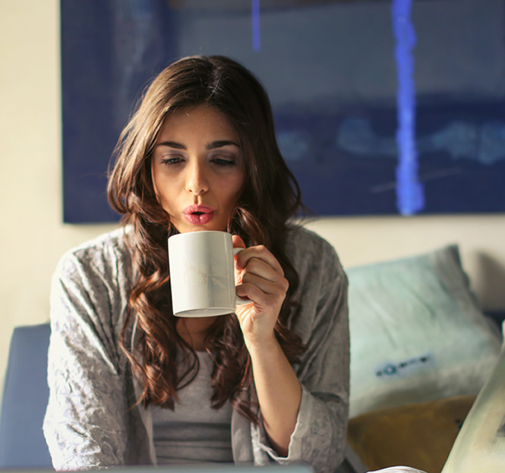 Woman blowing on a white mug, indoors. Wearing gray sweater, with dark hair and blue background.