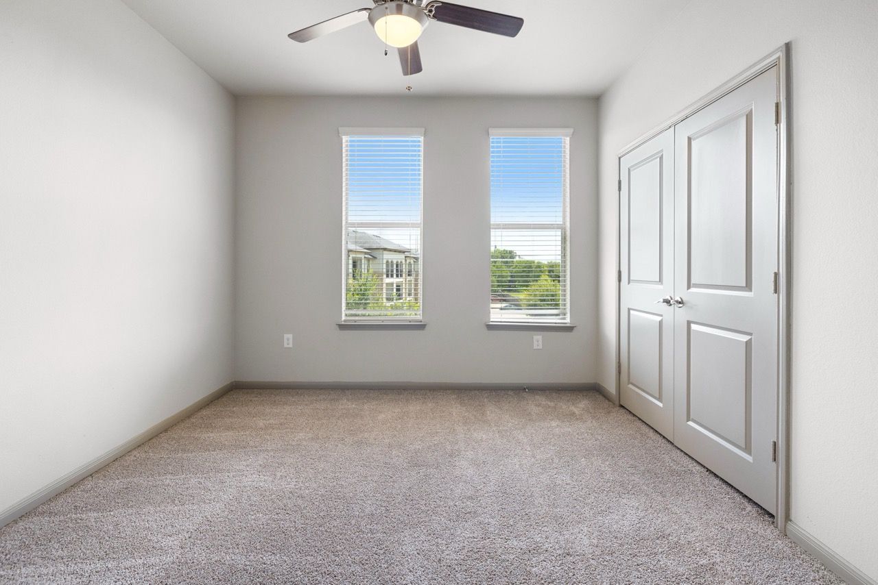 Empty bedroom with two large windows, a ceiling fan, beige carpet, and closet doors.
