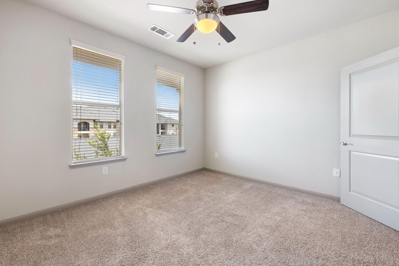 Empty bedroom with beige carpet, two windows with blinds, and a ceiling fan.