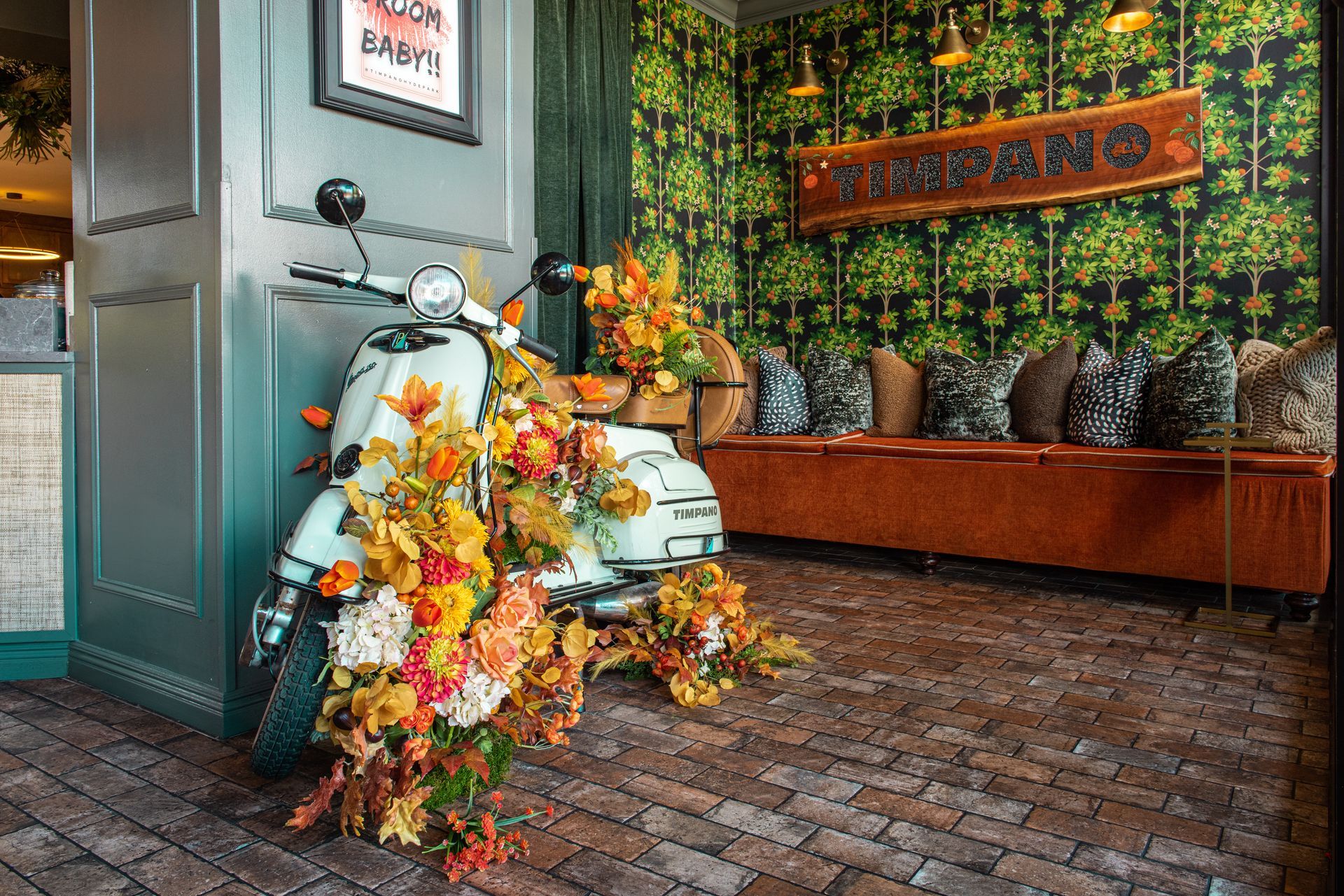 A cream-colored vintage scooter decorated with autumn flowers sits on a brick floor in a restaurant near a velvet bench.