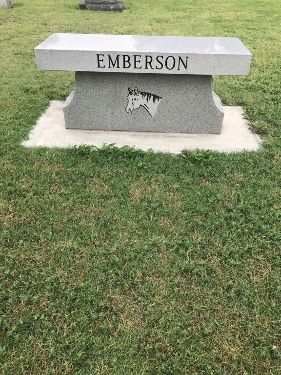 A graveyard bench with the name emberson on it is in the grass.
