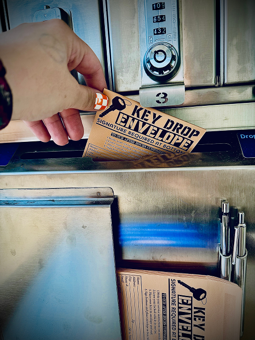 Hand dropping a key drop envelope into a metal drop box. The envelope is brown with a key graphic. | King's Auto LLC