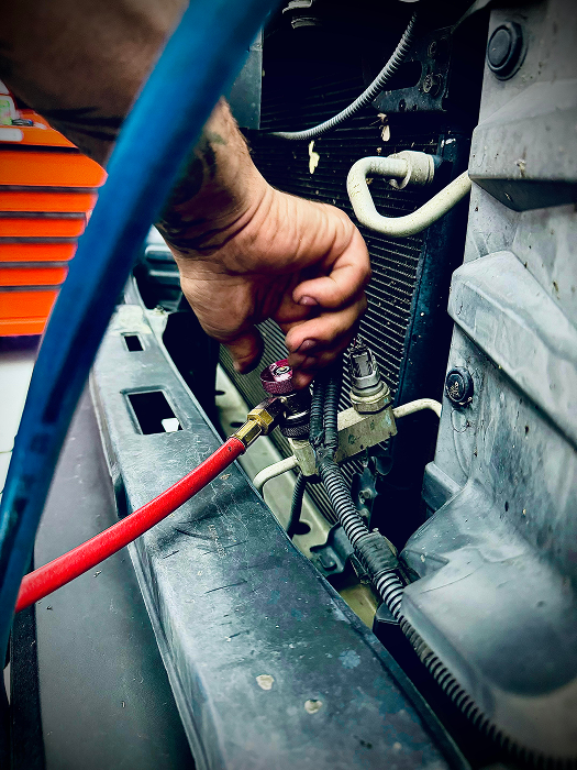 Mechanic connecting a red hose to a car's AC system near the grill, with a blue hose visible. | King's Auto LLC