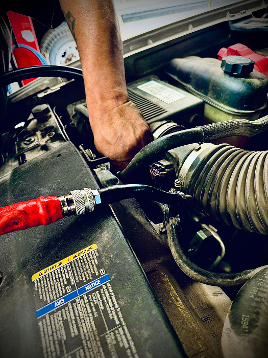 Person's hand working on a car engine. Black hose with red handle is visible. | King's Auto LLC