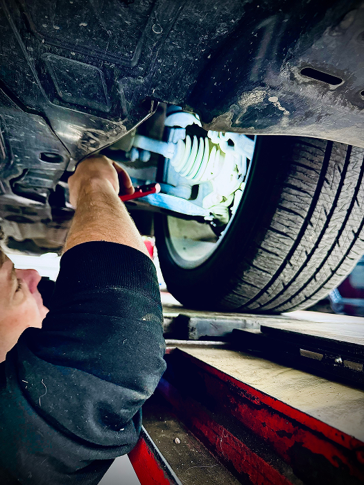 Mechanic working under a car, examining components near a tire, lit by a flashlight. | King's Auto LLC