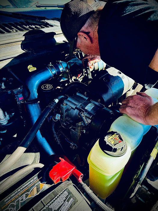Mechanic inspecting an engine, wearing a cap, over a blue intake. Coolant reservoir visible. | King's Auto LLC