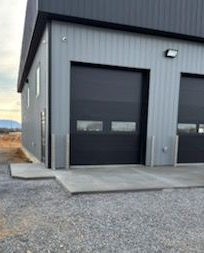 Gray industrial building with black garage doors, concrete pad, gravel.
