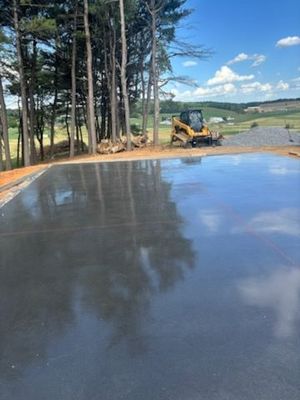 Newly poured, smooth, gray concrete slab reflects the sky and trees. A small yellow skid steer sits nearby.