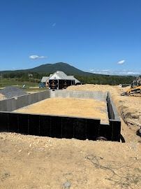 Construction site: foundation walls, dirt, sand, and mountain backdrop under a blue sky.