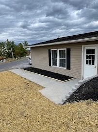 Tan building with black shutters and door, concrete path with steps, dark mulch, and cloudy sky.