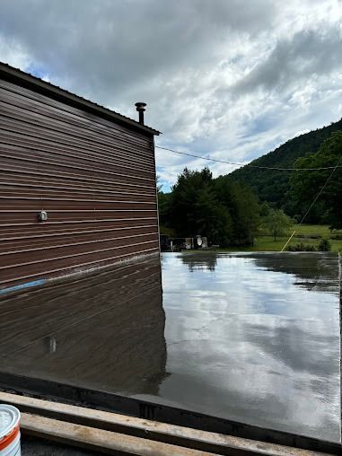 A freshly poured, wet concrete slab reflecting the sky, next to a brown-sided building with a mountainous backdrop.