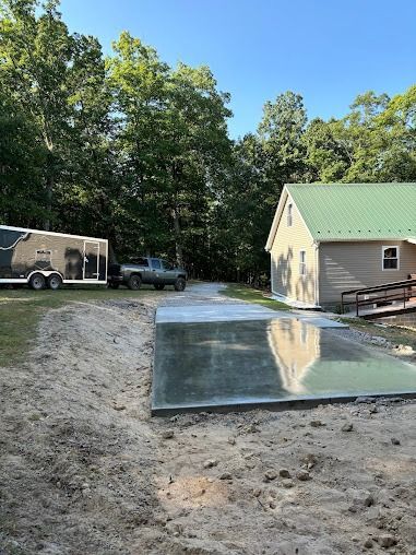 Concrete pad in front of building with green roof; truck and trailer on gravel driveway, trees in background.
