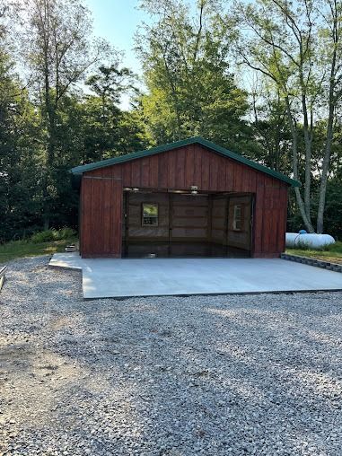 Red shed with an open front and concrete pad, on a gravel lot, trees in the background.
