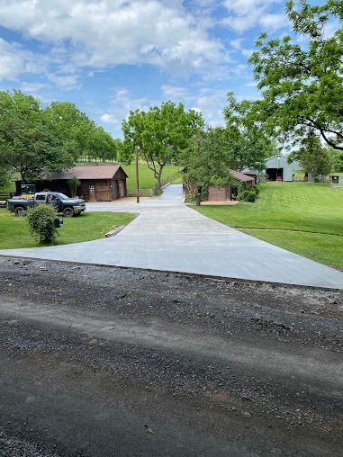 Gray concrete driveway leading to buildings and green trees under a blue sky.