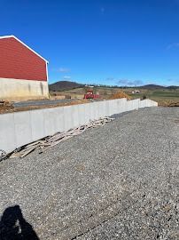 A concrete wall at a construction site with a red building and gravel ground. Blue sky in background.